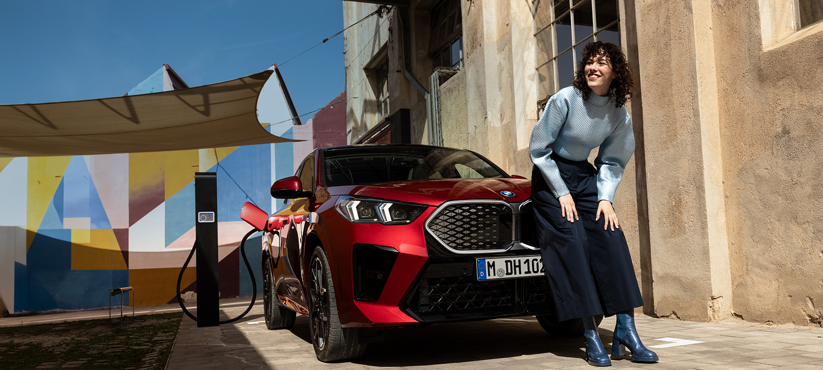 Eine Frau lehnt sich lachend gegen die Motorhaube eines roten, elektrischen BMWs.